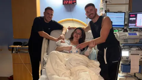 Family handout Krista, who is lying on a hospital bed, is holding Leven and smiling. Adam Frisby and Jamie Corbett are standing next to her. To the right is hospital equipment. 