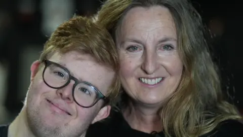 Jamie Niblock/BBC Edward Wagland with black glasses smiles at the camera while his head rests on a Helen Wagland's shoulder. She has shoulder length blonde hair with some grey. She is wearing a black top and smiling.