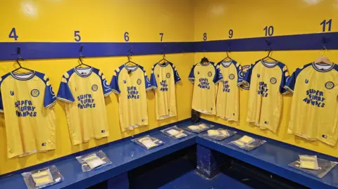 Matt Jarrett Team shirts hang in the men's changing rooms, yellow in colour with blue sleeves and blue writing on the front saying Super Furry Animals. 
