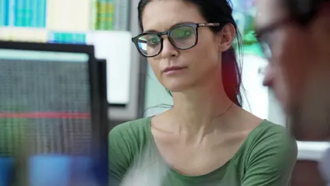 Getty Images Woman wearing glasses looks at computer screen . A man's blurred face is in the foreground and more sets of data are visible on a screen in the background