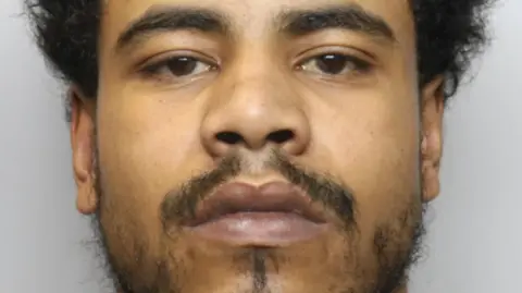 Greater Manchester Police A close-up mugshot of a young man with black frizzy hair, a sparse beard and dark eyes. He has a neutral expression.