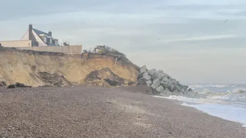 The Thorpeness coastline that has been eroded away. Large cliffs can be seen where the erosion has happened. Rock sea defences have been put around the cliff in the distance. A house above the cliff can be seen. 