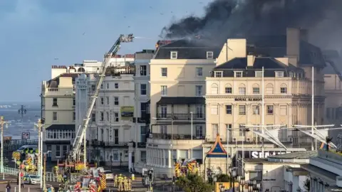 The Royal Albion hotel on Brighton seafront with black smoke coming from its roof and firefighters working to extinguish it.