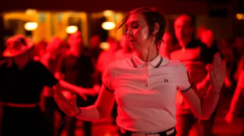 A woman with short brown hair and wearing a white polo shirt dances to Northern Soul music. She is bathed in red and white light and other dancers are visible in the background.
