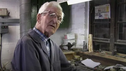 A man, Stan Shaw, with grey hair, wearing glasses and blue overalls in a workshop