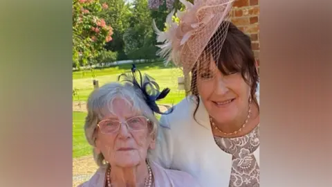 Beverley wears a smart white blazer, a beaded necklace and a pink fascinator hat. She has brown hair. On the left is her mother, wearing glasses, a pink dress or blazer, and appearing with white hair. It is sunny outside.