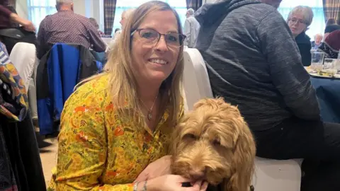 A woman with long blonde hair, wearing a yellow shirt and glasses, holds a golden-coloured dog in a busy dining room.