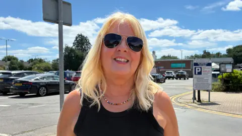 Zac Sherratt/BBC A woman with blonde hair wearing sunglasses and a black top stands in a car park, smiling into the camera.