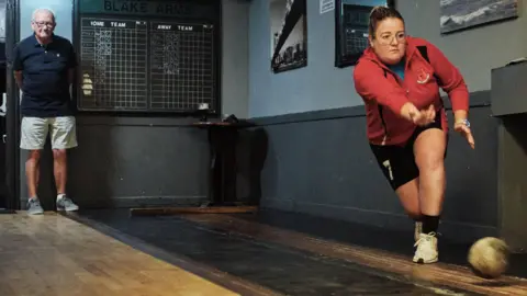 Joss Barratt A woman is shown throwing the ball down a skittle alley. She is wearing a red top, black shorts and white trainers. A man stands and watches in the background.