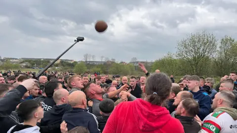 A large scrum made up of dozens of men is gathered on a field. A brown oval ball is in the air, with hands from players reaching out for it.