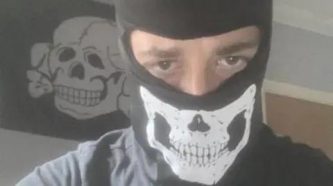 Alfie Coleman wearing a black balaclava with a skull on it. Only his eyes can be seen. Behind him a black flag with a white skull and crossbones is pinned to a wall.