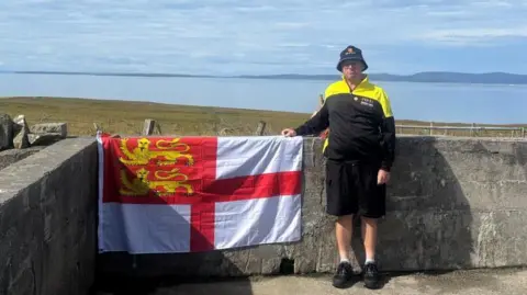 Kevin Adams is smiling as he stands next to the flag of Sark by the coast, with the sea in the background. He is wearing the yellow and black kit worn by the Sark team.