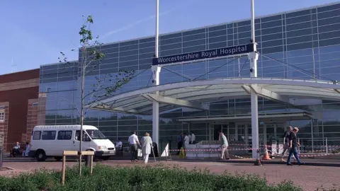 PA Media The exterior of Worcestershire Royal Hospital. It is a large glass-fronted building with a covered entrance. A white van is outside the building and a number of people are walking nearby.