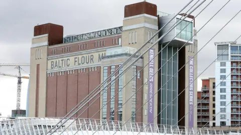 PA Media The Baltic Centre For Contemporary Art is a tall red-brick building on the River Tyne. Near the top, black bricks are laid in way that spells Baltic Flour Mills. The white Millennium Bridge is in the foreground of the photograph.