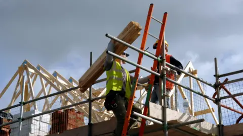 Two construction workers wearing hard hats and high visibility vests work at the top of a new house. There are scaffolding and timber beams.
