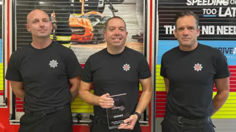 Three firefighters in matching black uniforms stand in front of a fire engine with safety campaign graphics on its shutter panels. The firefighter in the middle is holding an award plaque that reads “Winner.”