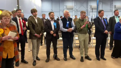 Image shows a row of people wearing different party rosettes with counting tables behind