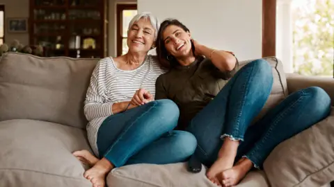 Getty Images Two generations of women sit on a sofa smiling and holding hands with a cabinet and picture in the background.