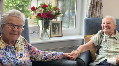 Jean Eyre and Terry Eyre. Jean has short white hair and glasses. She is smiling at the camera. Terry has white short hair and is looking at Jean and smiling. Both of them have their arms on the chair armrest and are holding hands. 