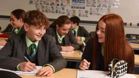 Pupils sat at their desks in a classroom at Walney School. Two pupils in the foreground, a boy and a girl, are looking at each other and smiling. Other pupils are writing and talking to each other. They all wear green shirts with black, blue and green ties and grey blazers.