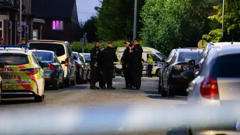 JMG Press A number of police officers in uniform stand between parked cars on a terraced street, with police tape across the road in the background and a police van