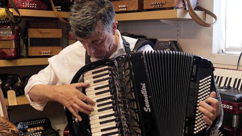 Rob Beecroft playing an accordion in his workshop with rows of instruments on shelves behind him