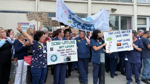 Nurses in navy scrubs and members of the public holding a protest outside a hospital building holding up home-made banners and signs reading things like 'tiny lives matter' and 'save our scbu'