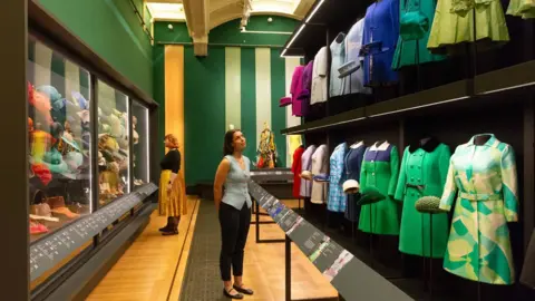 Royal Collection Trust Visitors look at colourful dresses and hats displayed on mannequins in the museum gallery, with glass cases of accessories on one side and information panels at knee height.