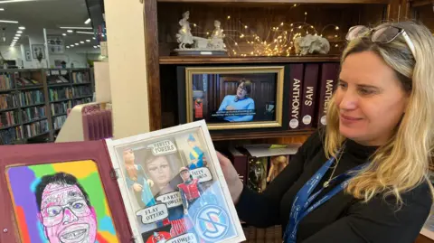 A picture of a blonde-haired woman, who is standing up. She is holding a book that features a self-portrait of a man and pictures of Gerry Anderson puppet characters.