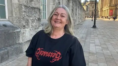 A woman with long hair in a black T-shirt with Cherrydead written in red letters on the front. She is stood outside on a brick pavement.