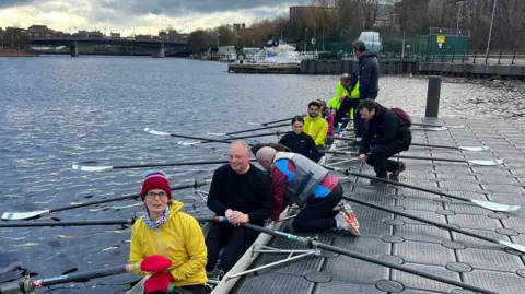 Tees Rowing Club Rowers board a rowing boat on a plastic pontoon on the River Tees. They are smiling and are wearing woolly hats and winter clothing. It looks early in the morning. 