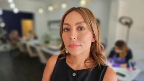 Lauren has light brown, shoulder length hair and is wearing a black sleeveless dress with brown buttons. She is standing in front of a long table where customers are having their nails done.