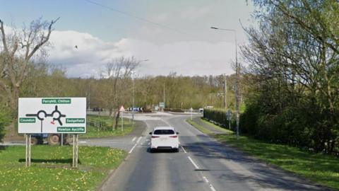 Streetview showing a white car approaching a roundabout next to a large roundabout sign