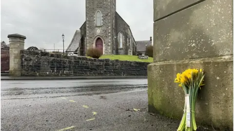 The side of a road with yellow flowers lying against a building. A church is the in background.
