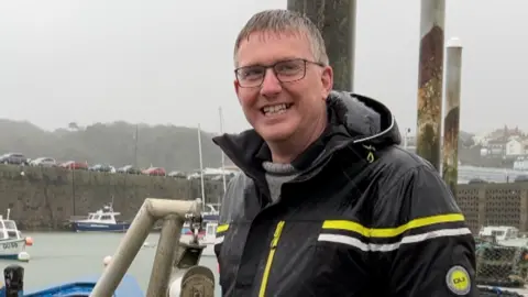 Man with short grey hair standing on a pontoon smiling at camera with rain water running down his face