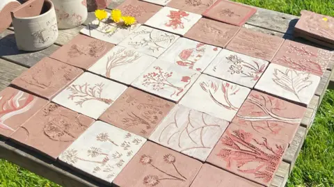 Leach Pottery A range of tiles with different designs including plants, flowers and houses can be seen on a table. Three dandelions can be seen on the left. The table is on grass.