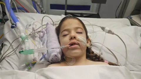 Marie Wood A young girl, asleep in her hospital bed, with ventilator, drips and various other wires and hospital equipment around her.