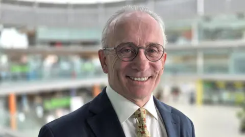 David Bick is smiling. He's wearing a navy suit jacket over a white shirt and patterned tie. The inside of the Norwich Forum is behind him but out of focus.