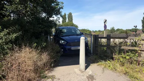 Jamie Downsworth A blue car parked between two railings on a footpath, behind a concrete bollard. Large trees and bushes can be seen on the left. There is a wooden fence on the right. The sky is blue.