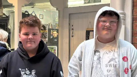 BBC/Eve Connor Monty and Joel. Monty is wearing a black hoodie with a cartoon design and has brown hair. Joel is wearing a white t-shirt with Taylor Swift on, and a grey zip hoodie. Joel also has glasses on, and has brown hair. They are standing outside Regency Records.