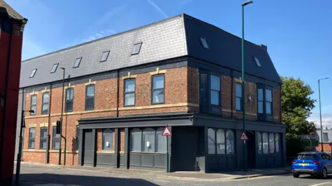 The three-storey brick building is on the corner of the street. It has black exterior panelling and multiple rectangular windows on the upper floors and arched widows on the most lower.