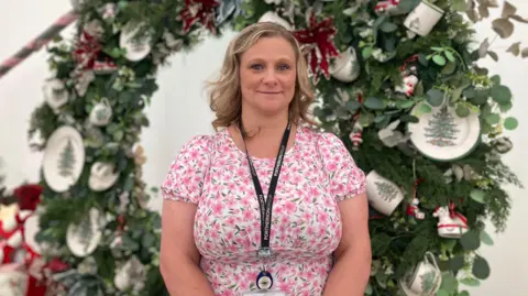 A woman with wavy hair in a pink floral top is looking at the camera. Behind her is a festive display including plates with Christmas trees on 