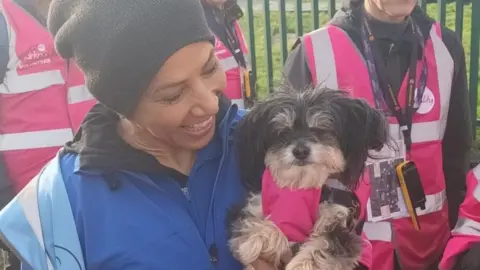 Dame Kelly Holmes smiles and looks at her dog, a Shih Tzu type breed, which is wearing a little pink hi-vis top. She is wearing a blue coat, a grey beanie, and a blue hi-vis tabard. There are other runners wearing pink hi-vis tabards standing in the background.