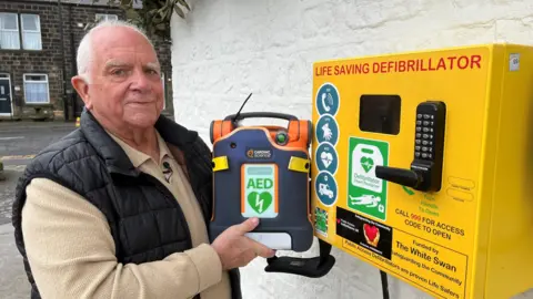 A man in his late 70s is stood next to a bright yellow box that is used to store a public access defibrillator. He is holding up one of the life-saving devices in his hands.