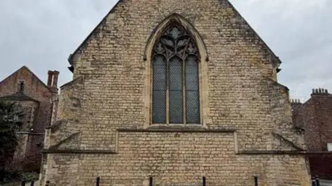 St John University/City of York Council The end wall of a stone built chapel. The wall forms a large triangular gable rising to a peak at the top center. A large central window contains three vertical lancet sections topped by decorative stone tracery.