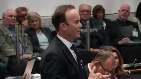 John Fairhall/BBC Richard Rout is wearing a black suit and tie with a white shirt. He is standing in the council chamber. He is speaking and is animated. 