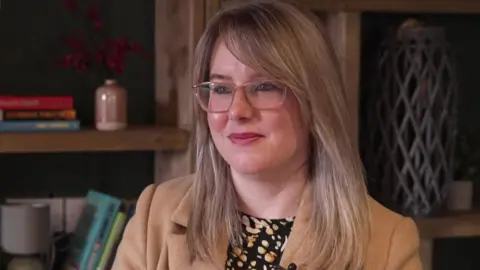 Kirsty Walsh - The image shows a woman seated indoors against a backdrop of wooden shelving units. The shelves are filled with a mix of books, decorative items, and small ornaments. Some of the items include stacked books, a ceramic jug, a small vase, and a tall lattice-style decorative object. The woman is wearing a light brown or camel-coloured coat over a patterned top featuring black and pale shapes. 