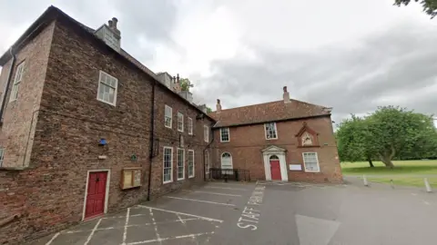 Google An L-shaped brick-built house, dating from the 1730s, has red doors and white window frames. It stands in a yard used for a car park, with a green field and trees in the background.