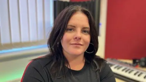 Alex Bish/BBC A woman with long dark hair, a grey top, and hoop earrings is sitting in a production studio at the Catch 22 office in Farnham, there is a keyboard on the table behind her