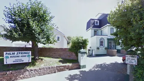 Former Palm Springs Nursing Home in St Helier, showing the entrance driveway, roadside signs, mature trees and the main building before its closure in 2021.